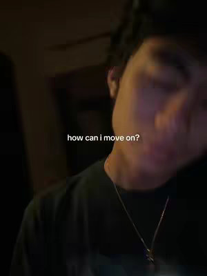 A dimly lit selfie focuses on a young person's face, slightly out of focus. The person, wearing a dark shirt and a subtle chain, tilts their head, with text overlayed asking "how can I move on?". The background is dark and indistinct.