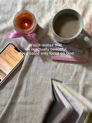 An overhead shot of items on a white, slightly wrinkled surface. These items include a lit candle in a glass jar with a pink ribbon, a smartphone displaying text, a mug of coffee with a spoon, two pink and pale purple rectangular objects, and an open book. The background is a soft, patterned white fabric. Text is overlaid on the image.