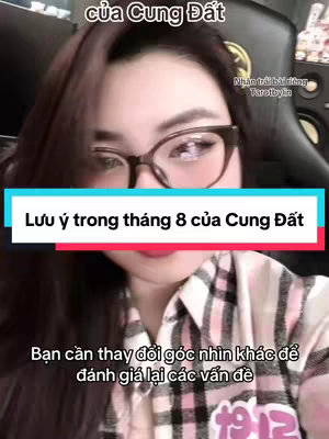 A selfie-style video cover with a woman at the forefront. She is wearing glasses, and the frame is mostly close up on her face and upper body. A rectangular bar with the text "Lưu ý trong tháng 8 của Cung Đất" is placed over her image. She is wearing a plaid shirt and the background is blurred. The overall color scheme is quite warm, thanks to the lighting. The composition places the text with a prominent placement in the image's center with the woman's face partially covering the text.
