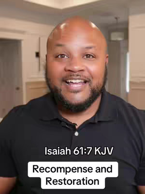 A close-up shot of a Black man with a bald head and a neatly trimmed beard. He is smiling with his mouth open, and the background is slightly blurred but appears to be a well-lit interior with white walls and a light fixture.