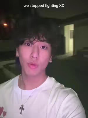A young man with dark hair and a white t-shirt is centered in the frame, facing the camera with an expression that could suggest a moment of relief or a shared inside joke. Above him, overlay text reads "we stopped fighting XD", hinting at a narrative of reconciliation or amusement. The background is blurred, suggesting a night scene or a dimly lit environment. The composition is simple, focusing primarily on the man's face and expression, with the text overlay providing context to the image.
