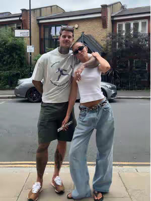 A photograph of a couple posing outdoors. They are standing on a sidewalk with a building and parked car behind them. The man on the left is wearing a white t-shirt, shorts, and sneakers. He is holding sunglasses on top of his head. The woman on the right side wears a white crop top, light blue jeans, and sunglasses. They are embracing with each other.