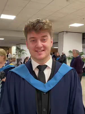 A young man in a graduation gown stands in a brightly lit hallway. He's smiling warmly at the camera, a blue and black graduation stole over his shoulders and a black tie and suit visible beneath the gown. Other people stand in the background, and the composition is a medium shot.