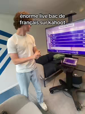 A young man, wearing a white t-shirt and light-blue jeans, stands facing left in a room with a large-screen TV and a laptop on a rolling stand. Text overlayed on the image reads, "énorme live bac de français sur kahoot."
