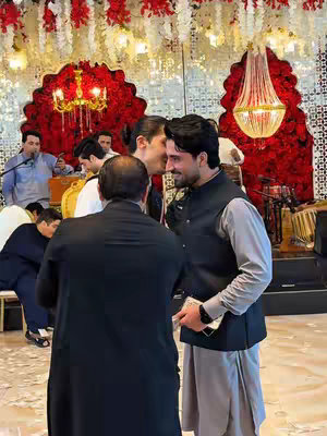 The image captures a social gathering, likely a wedding or celebration, with several individuals interacting. The scene is brightly lit, with a focus on individuals in the foreground, with many in the background and with multiple people in nice attire. A red decorative backdrop with floral elements is visible. The composition suggests an event of joy and celebration.