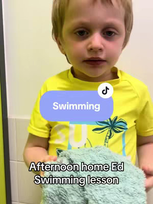 A close-up shot of a young boy with blond hair, wearing a yellow shirt with a palm tree design, holding a green towel. The background is a plain white wall. Text overlays at the bottom say "Afternoon home Ed Swimming lesson".