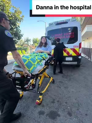 A woman is being transported on a stretcher towards an ambulance. She is lying on a stretcher covered with a patterned blanket. The ambulance is in the background, and two people are assisting the stretcher. There is text overlayed on top.