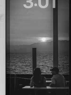A black and white photo through a window, of a couple seated at a table with their backs to the viewer, watching the sunset over a lake. The timestamp '3.01' is visible at the top of the image, the sun, and the silhouette of mountains in the distance.