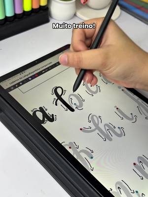 A tablet displaying calligraphy practice, with a hand holding a stylus, the phrase "Muito treino!" is added as text. There are various stationary items in the background, as well.