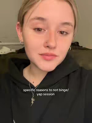 A close-up selfie of a person with short, light-colored hair, wearing a black hoodie. The person is looking towards the camera with a neutral expression. Text overlaid at the bottom reads 'specific reasons to not binge/yap session'. The background is a soft, out-of-focus domestic setting.