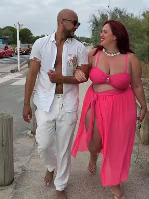 A couple is walking together by the beach. The woman on the right is smiling and looking towards the man, the man is wearing glasses and looking at the woman. They are both walking next to each other and holding hands as if a moment of intimacy.