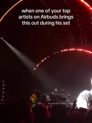 A stage with a DJ setup is lit with red and white lights. A spotlight focuses on the center. Text overlayed reads 'when one of your top artists on Airbuds brings this out during his set'.