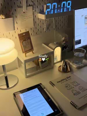 An aesthetic study desk setup is shown with a digital clock showing '20:09', an iPad displaying what looks like notes, multiple pens, a book titled 'Healthy diet', and a small white shelf with a light and decorative items. The lighting is warm, creating a cozy atmosphere.