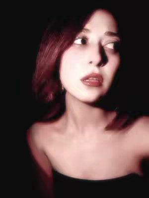 A portrait of a woman with red hair against a dark background. She is looking to the side with a thoughtful expression. The image has a soft, slightly blurred focus, creating a dreamy or ethereal effect.