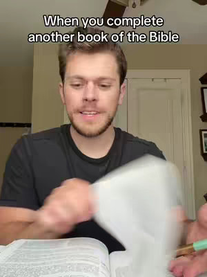 A man with short brown hair is holding open a Bible and flipping through the pages. The text overlay reads, "When you complete another book of the Bible". The man is wearing a black t-shirt. The background includes a ceiling fan and neutral-colored walls and trim.