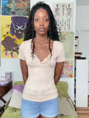 A young woman stands facing the camera, with a neutral expression. Some background elements, including artwork on the wall. She wears a beige top and denim shorts.