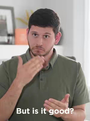 A man with short dark hair and a short beard is looking down with a serious expression. He is wearing a green polo shirt. His right hand is touching his throat, and his left hand is held up to his chest with the palm facing out. The text overlay 'But is it good?' is positioned at the bottom.