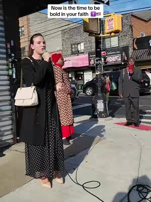 A woman is standing on a sidewalk speaking into a microphone. She is wearing a black coat over a long black dress with white polka dots, and is carrying a white purse. Behind her are two other people and a street scene with buildings and traffic lights. Text is overlayed on the screen.