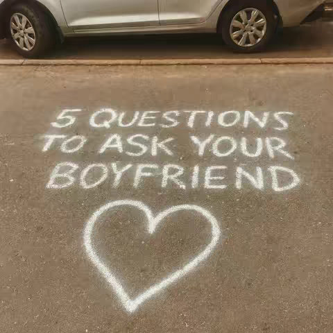 An overhead shot shows a section of a gray or brown pavement with a silver car parked at the top edge of the frame. The words '5 QUESTIONS TO ASK YOUR BOYFRIEND' are written in large, white chalk on the pavement. Beneath the text is a simple heart drawn with the same white chalk.