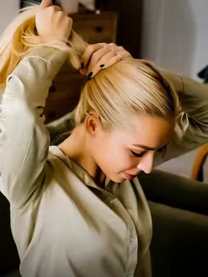 A woman with blonde hair pulls her hair up into a ponytail with her hands. She wears a light green button-up shirt, and part of a dark green sofa and a wooden chest are visible in the background.