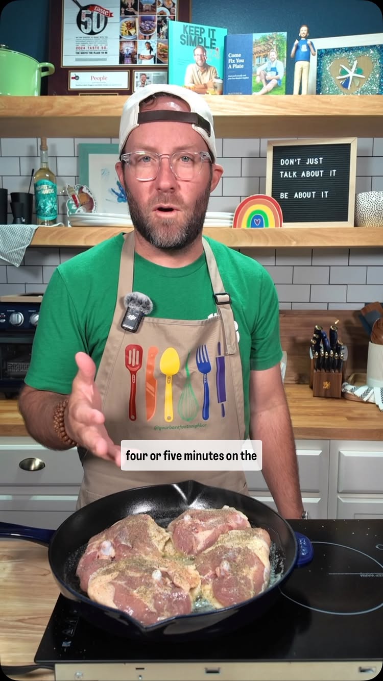 A man in a green shirt and apron stands in a kitchen, gesturing toward a pan of chicken thighs on a stovetop. A banner with text is visible, along with images and books on the wall behind him. The man is smiling and looking at the camera, and the photo is framed with a focus on the cooking process, and has some food-related text and a stove in the foreground.