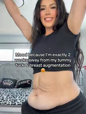A woman is smiling, arms raised above her head, in front of a bed. Text overlay indicates she is excited about upcoming surgery. The focus is on the midriff showing the stomach, which is a key part of the video's content. The surrounding environment includes a bed with decorative pillows, suggesting a bedroom setting.
