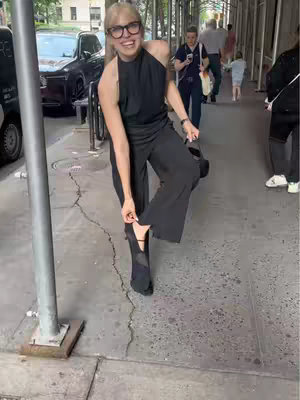 A woman in black clothing and glasses stands on a sidewalk in New York City. She is adjusting the strap of her shoe, with a look of surprise and excitement. The background includes other people, buildings, and scaffolding.
