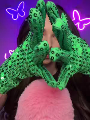 A person is centered in a vertical image, with green sequined gloves forming a heart shape in front of their face. The top half of the image is a dark purple background with neon butterfly outlines, and a fluffy pink cushion is in the bottom half. The person's eyes are not visible due to the angle and the gloves.