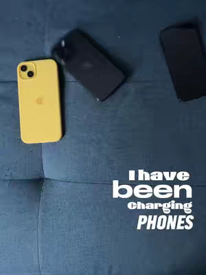 The image shows three smartphones on a textured blue surface, possibly a couch. One phone has a yellow case, is on the left side, and is oriented with the camera at the top. Another phone is to the right, black in color. The third phone is black, and is in the center. Prominently displayed in the lower-right corner is white text that says, "I have been charging PHONES".