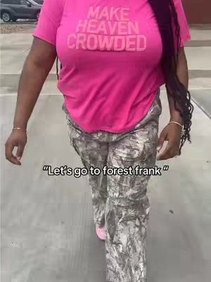 A woman wearing a bright pink shirt with the words "MAKE HEAVEN CROWDED" printed on it and camouflage pants is walking on a sidewalk towards the camera.