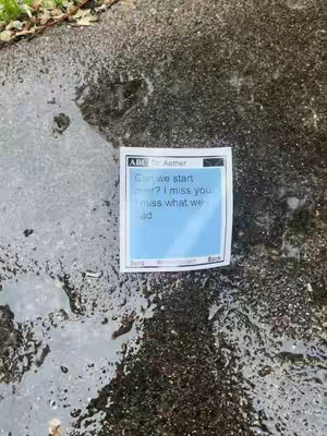 A square, blue sticky note with a text message on it is laid on a wet, textured surface. The surface appears to be a dark, rough pavement with water droplets reflecting the surrounding environment. The note is positioned directly on the surface with a white border. Text on the note reads 'Can we start over? I miss you miss what we had'. The overall composition has a somber tone, emphasizing the solitary note against a cold, wet background.