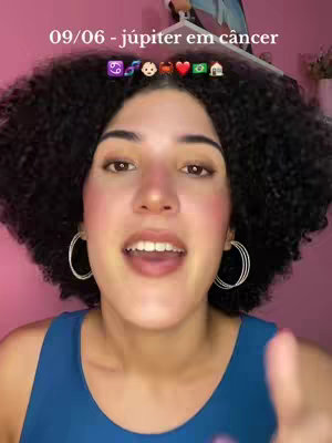 A young woman with curly dark hair and hoop earrings is centered in the frame, smiling and gesturing with her right hand. The background is a soft pink, and text overlays include the date and astrological symbols.