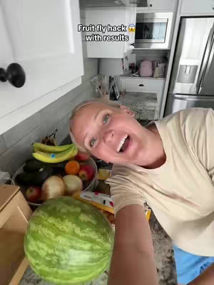 A woman with blonde hair smiles wide, leaning toward the camera in a bright kitchen. A watermelon is held out in the foreground, and a fruit bowl stands behind her. Text appears at the top of the frame.