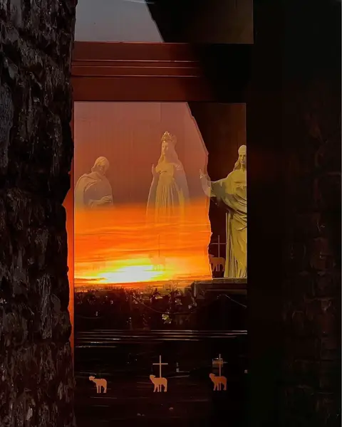 The image captures a religious scene viewed through a window. It depicts a vibrant sunset or sunrise painting the sky with orange and yellow hues. Through the window frame appear statues, possibly of religious figures. The foreground is partially obscured by the dark framing of the window, and the scene is framed on the left by a dark stone wall.