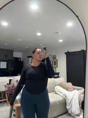 A woman is taking a selfie in front of a large, arched mirror. She's wearing a black long-sleeved top and dark blue leggings. The background includes a beige couch, artwork on the wall, and a glimpse of a kitchen area.