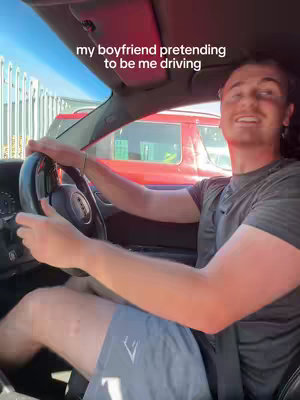 A man is driving a car. The man is smiling at the camera. Text overlay says: 'my boyfriend pretending to be me driving'.