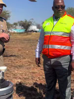 The image shows two men standing outdoors on a sunny day. One man is wearing a police-style uniform and cap, and the other is wearing a high-visibility vest over a shirt and pants.  In the background, there's dry grass, a white van, and trees. The overall shot is well-lit and appears to be taken in an open area.