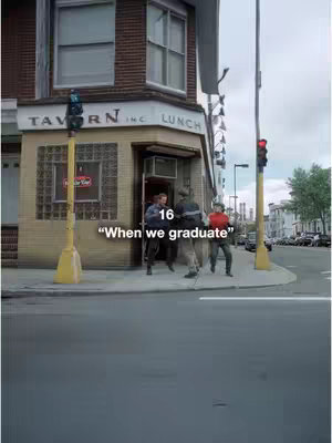The image shows the corner of a building with a sign that says Tavern inc. Lunch. There are three people on the sidewalk in front of the building. One person is wearing a red shirt. There are also some traffic lights and a street.