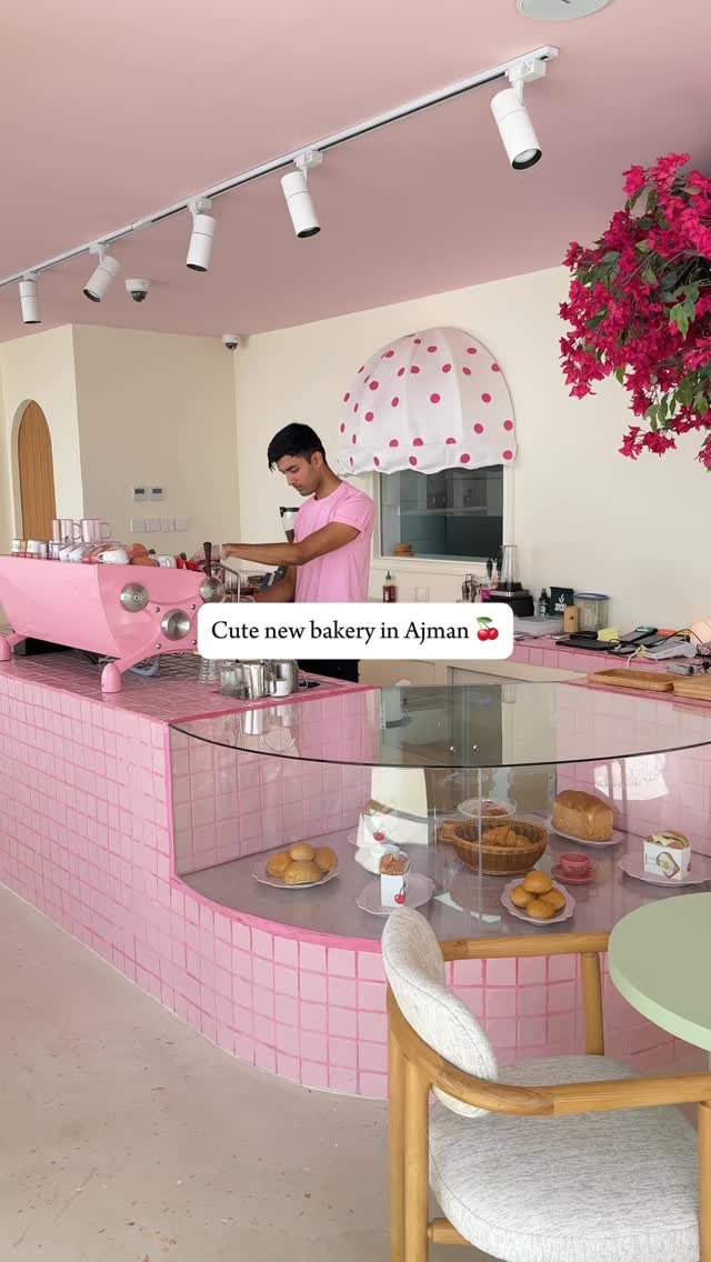 The image showcases a modern bakery interior with a man behind a pink counter preparing food. The space has a light pink ceiling, white walls, and spotlights. A pastry display case is visible, filled with baked goods. The man is wearing a pink shirt. Additional elements include a table, chair, and decorative plants, all contributing to a cute and inviting atmosphere.