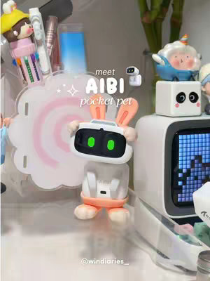 The image features a desk setup with various items. The focal point is a small robot with rabbit ears, a white body, and a black screen. Around it are a white cloud-shaped stand, pens, and a small pixel art display with a winking face. To the right, there’s a tall green plant. The background is slightly blurred, with a vibrant color palette.