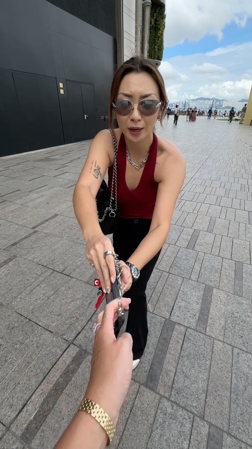 A woman in sunglasses and a red top stands outside, extending her hand towards the camera. She holds a phone attached to several charms. The setting is an urban environment, with buildings and pavement visible, implying a city backdrop. A person is holding a phone in their other hand, which is about to touch the phone in the other woman's hand.