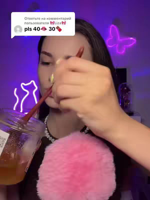 A woman is holding a jar with a honey-like substance, with a pink fluffy object in the foreground. The background has neon lights shaped like a butterfly and cat. Text overlay is present.