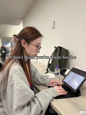 The image shows a young woman with glasses working on a laptop at a desk. Text overlayed says, 'I don't think you're ready for the'. The setting is a brightly lit room. In the background, a couple of people are walking.
