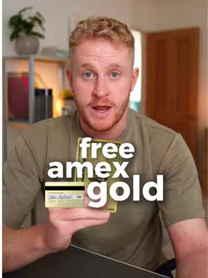 A man with short, reddish-blond hair and a slight beard holds up two credit cards in front of him with the text overlay "free amex gold". He is wearing a solid-color olive green t-shirt. The background is blurred, but part of a wooden door and shelving unit are visible.