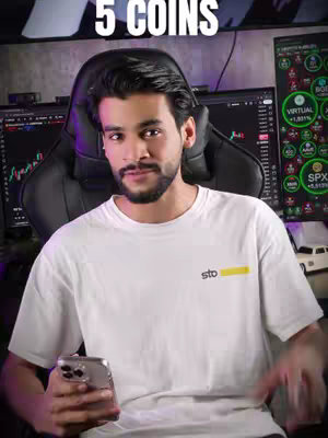 A man with short black hair and a beard is the central focus of the image. He's wearing a white t-shirt with a yellow and black logo. He is seated with a phone in hand, positioned in front of two computer screens displaying financial data charts. The background features a black office chair and dim lighting with the text 5 COINS overlaid at the top.