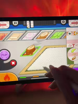 The image shows a person's hand holding a stylus, interacting with a tablet screen displaying a cooking simulation game. The gameplay appears to involve food preparation, with various ingredients lined up at the top and a cooking area in the center. The color palette is dominated by the colors used in the game, along with red, which seems to be from an external light source.
