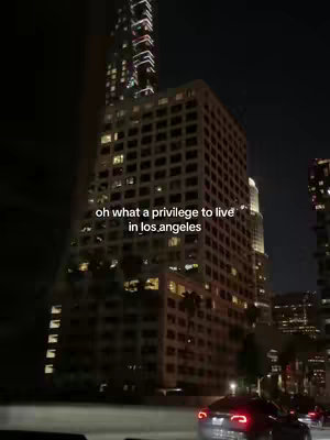 A night scene featuring a tall skyscraper in the foreground with lights on in the windows, with other buildings in the background. The shot is taken from the perspective of a moving vehicle, with car headlights visible in the bottom of the frame, and the text overlayed.