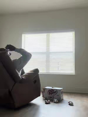 The image shows a person reclining in a brown armchair, with their arms resting overhead, possibly in relaxation. A box of Dr. Pepper cans and several loose cans are on the floor in front of the recliner, next to a window covered by blinds. The room has a minimalistic design with a light color palette.
