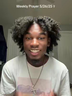 A young Black man with a curly hairstyle, wearing a white t-shirt and a silver cross necklace, smiles directly at the camera. The background is a neutral-toned room setting.