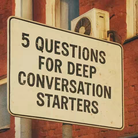 A slightly angled, close-up shot of a white sign with black text against a blurred background with red bricks, an air conditioning unit, and other architectural elements. The sign reads, '5 QUESTIONS FOR DEEP CONVERSATION STARTERS'.